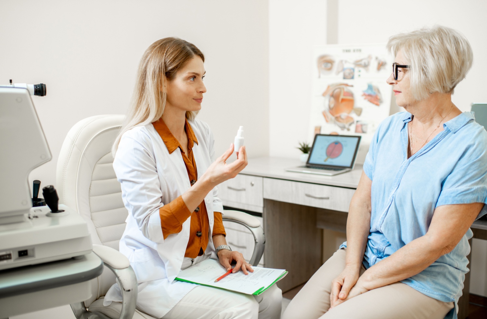 An optometrist holding a bottle of dilating eye drops and explaining to an older patient what to expect during their eye exam.